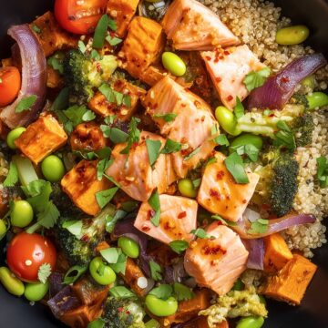 Overhead hot honey salmon sweet potato bowl with quinoa, roasted sweet potatoes, broccoli, edamame, cherry tomatoes, red onion, and fresh herbs, topped with chunks of glazed salmon in a matte black bowl on a rustic wood table.