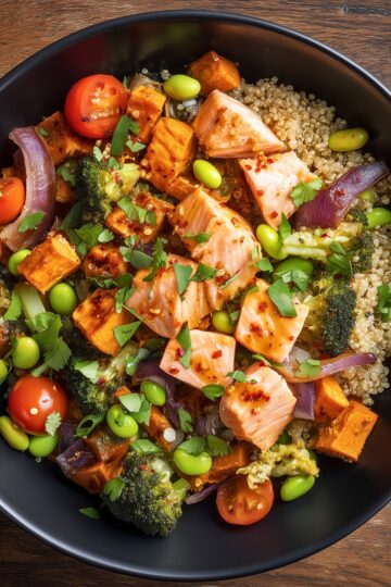 Overhead hot honey salmon sweet potato bowl with quinoa, roasted sweet potatoes, broccoli, edamame, cherry tomatoes, red onion, and fresh herbs, topped with chunks of glazed salmon in a matte black bowl on a rustic wood table.