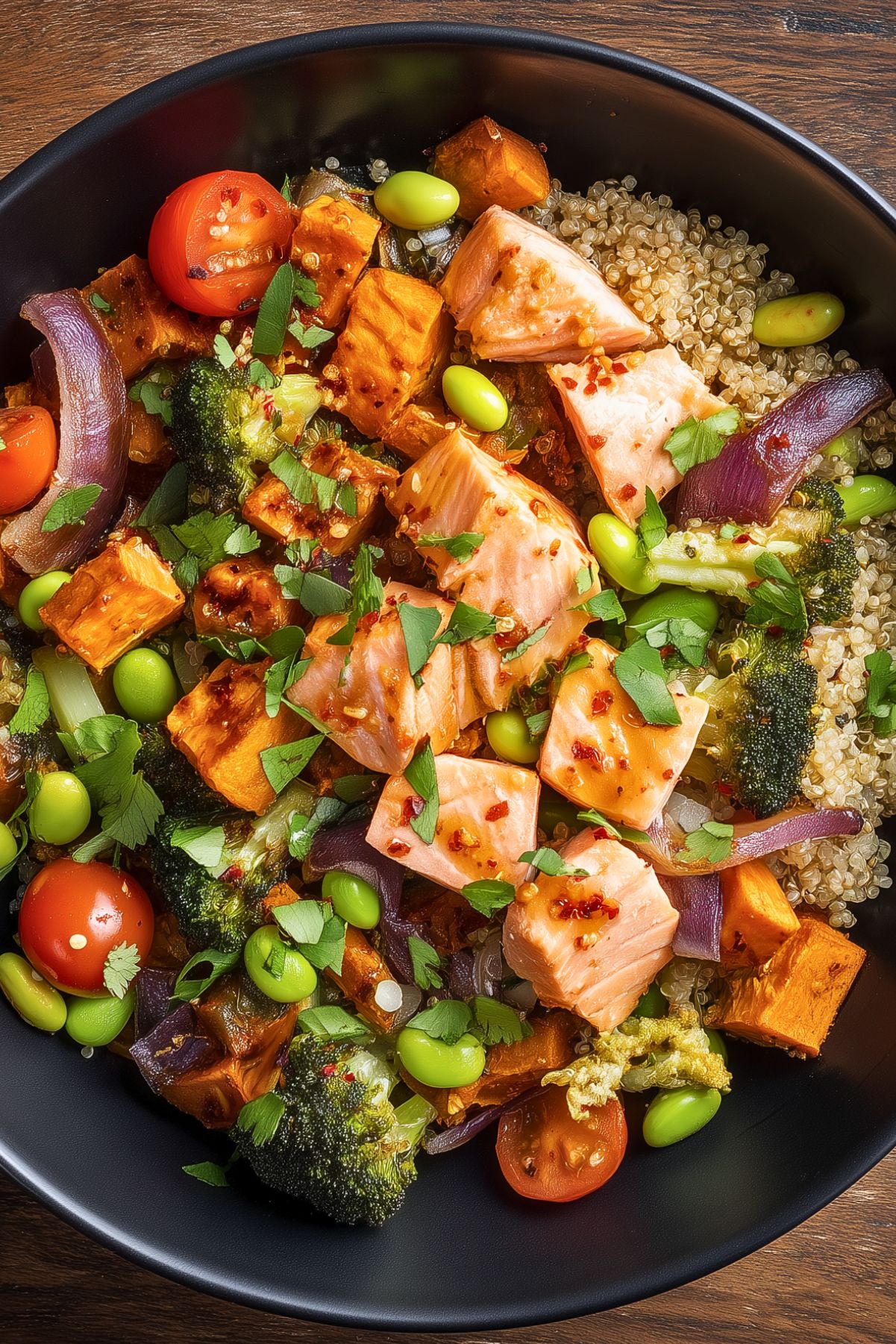 Overhead hot honey salmon sweet potato bowl with quinoa, roasted sweet potatoes, broccoli, edamame, cherry tomatoes, red onion, and fresh herbs, topped with chunks of glazed salmon in a matte black bowl on a rustic wood table.
