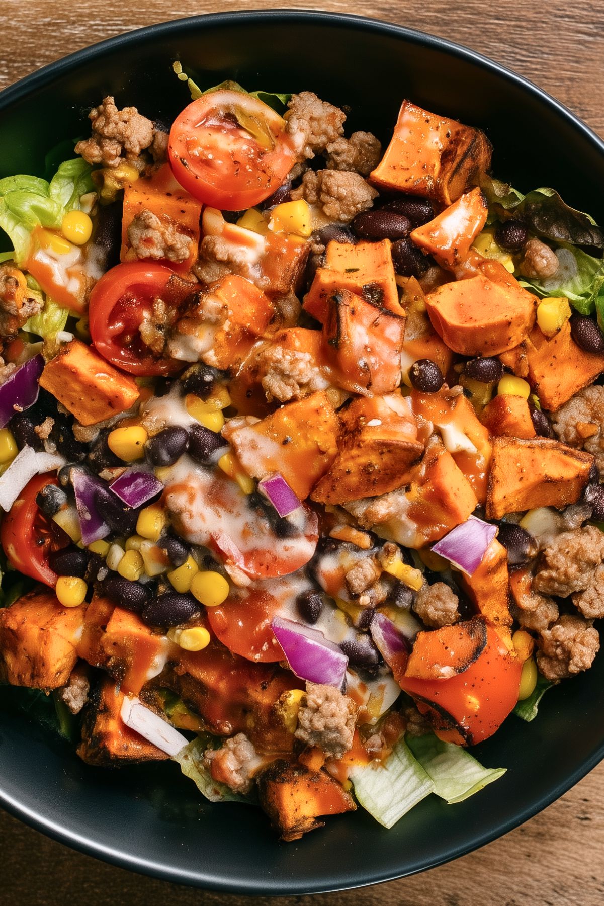 High protein hot honey turkey sweet potato bowl with ground turkey, roasted sweet potatoes, black beans, corn, tomatoes, red onion, lettuce, and creamy sauce in a black bowl.