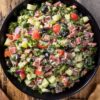 Overhead view of Italian sub grinder chopped salad with chopped deli meats, romaine, cucumber, tomatoes, red onion, black olives, and creamy dressing in a black bowl, with pita chips on a rustic wood table.
