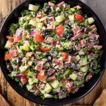 Overhead view of Italian sub grinder chopped salad with chopped deli meats, romaine, cucumber, tomatoes, red onion, black olives, and creamy dressing in a black bowl, with pita chips on a rustic wood table.