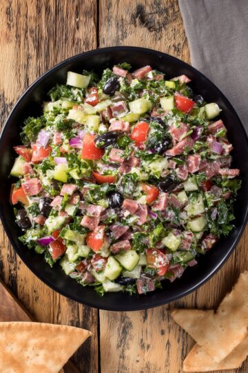 Overhead view of Italian sub grinder chopped salad with chopped deli meats, romaine, cucumber, tomatoes, red onion, black olives, and creamy dressing in a black bowl, with pita chips on a rustic wood table.