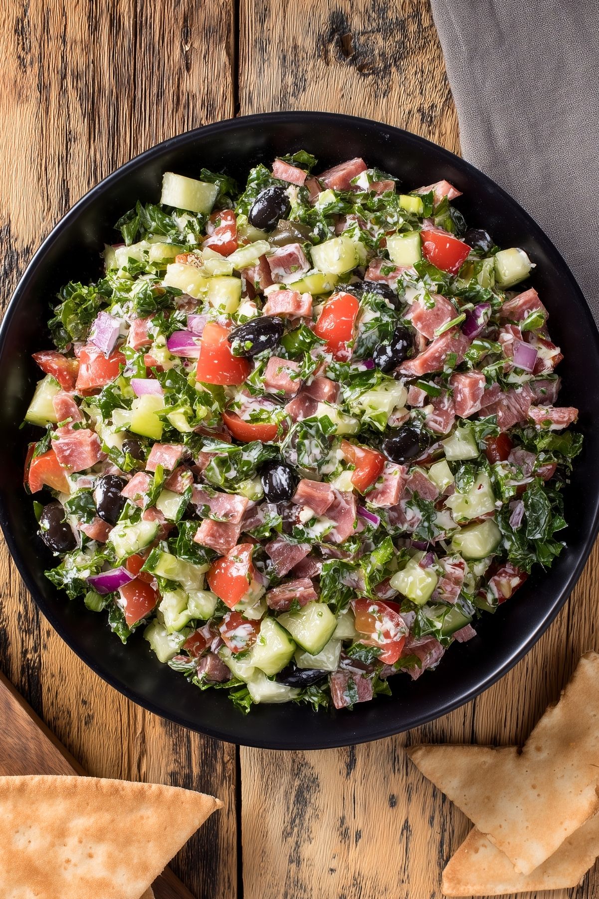 Overhead view of Italian sub grinder chopped salad with chopped deli meats, romaine, cucumber, tomatoes, red onion, black olives, and creamy dressing in a black bowl, with pita chips on a rustic wood table.