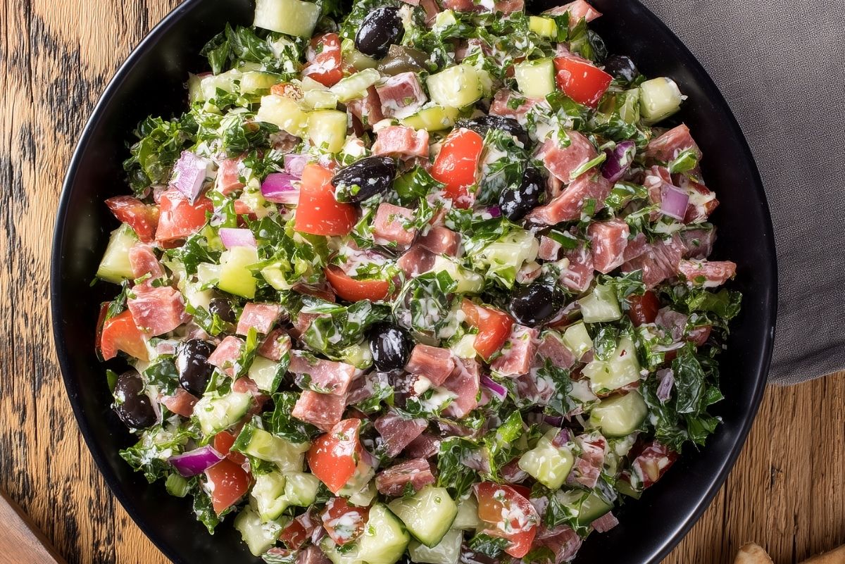 Close-up overhead view of Italian sub grinder chopped salad with deli meats, romaine, cucumber, tomatoes, red onion, black olives, and creamy dressing in a black bowl on rustic wood.