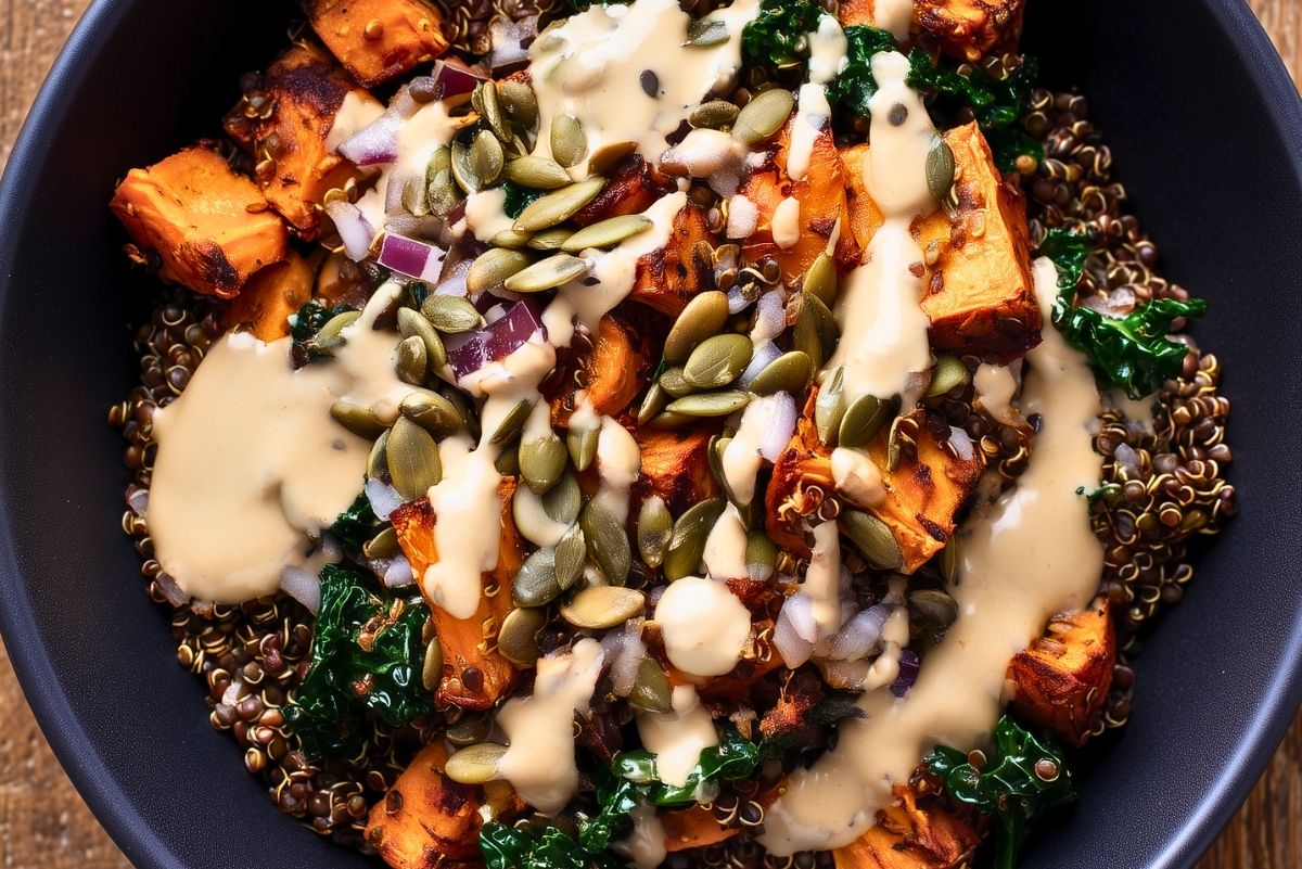Close-up of a lentil sweet potato protein bowl with roasted sweet potatoes, lentils, kale, red onion, pumpkin seeds, and creamy dressing in a black bowl.