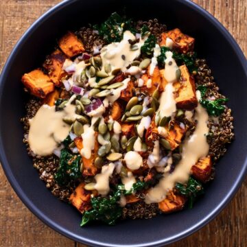 Overhead view of a lentil sweet potato protein bowl with roasted sweet potatoes, lentils, kale, red onion, pumpkin seeds, and creamy dressing in a black bowl on rustic wood.