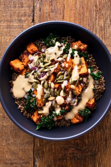 Overhead view of a lentil sweet potato protein bowl with roasted sweet potatoes, lentils, kale, red onion, pumpkin seeds, and creamy dressing in a black bowl on rustic wood.
