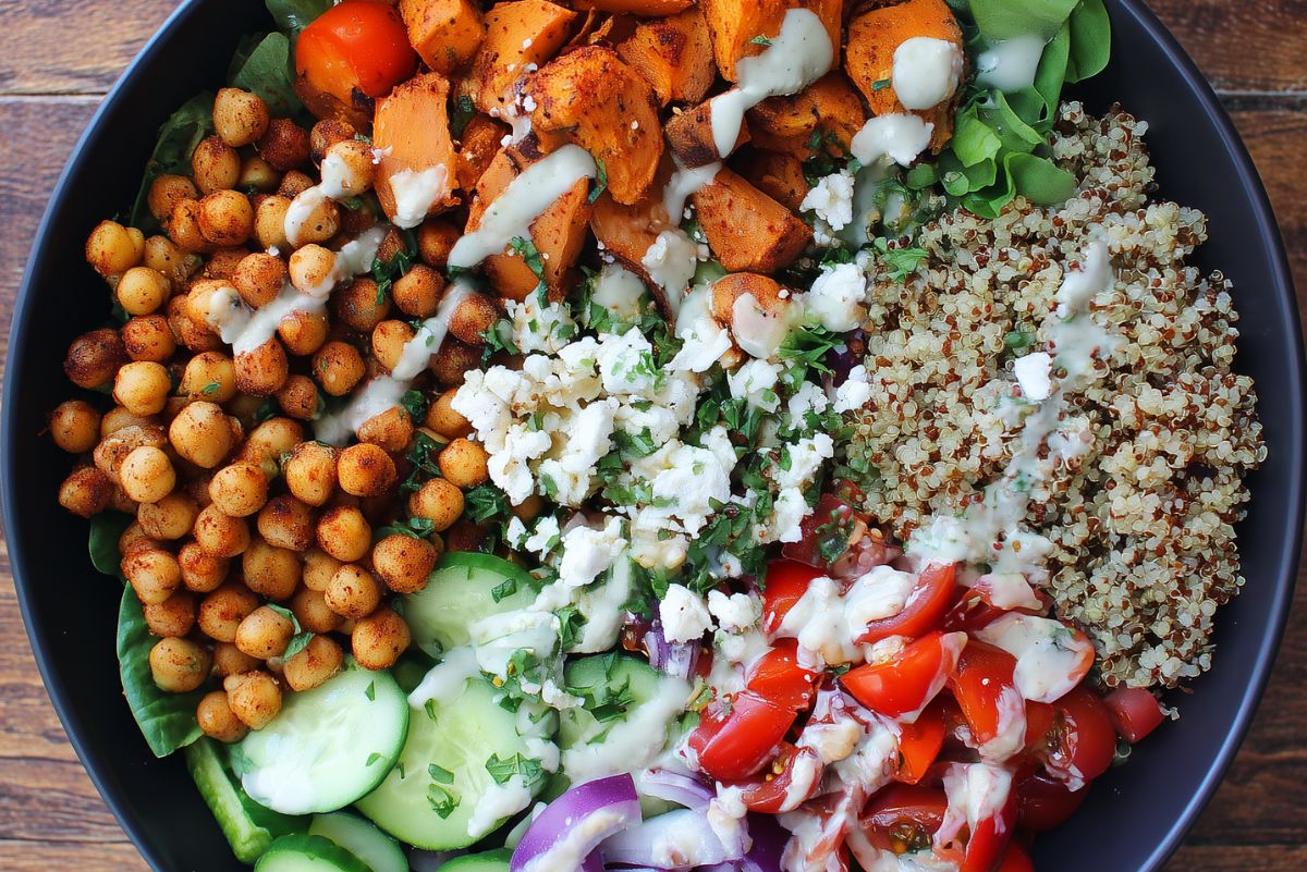 Close-up Mediterranean chickpea sweet potato bowl with quinoa, roasted chickpeas, seasoned sweet potatoes, cucumbers, tomatoes, red onion, greens, feta, herbs, and creamy dressing.