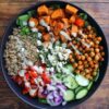 Overhead Mediterranean chickpea sweet potato bowl with quinoa, roasted chickpeas, seasoned sweet potatoes, cucumbers, tomatoes, red onion, greens, feta, herbs, and creamy dressing.