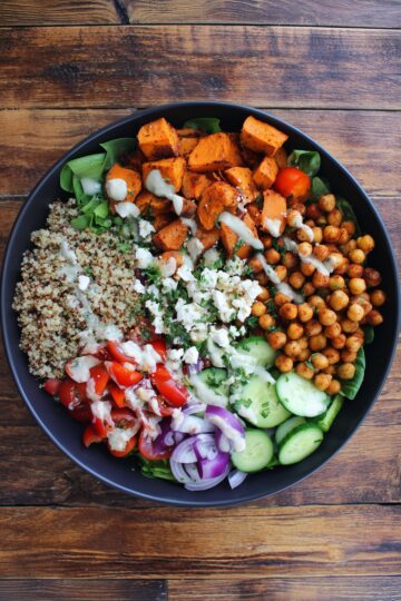 Overhead Mediterranean chickpea sweet potato bowl with quinoa, roasted chickpeas, seasoned sweet potatoes, cucumbers, tomatoes, red onion, greens, feta, herbs, and creamy dressing.