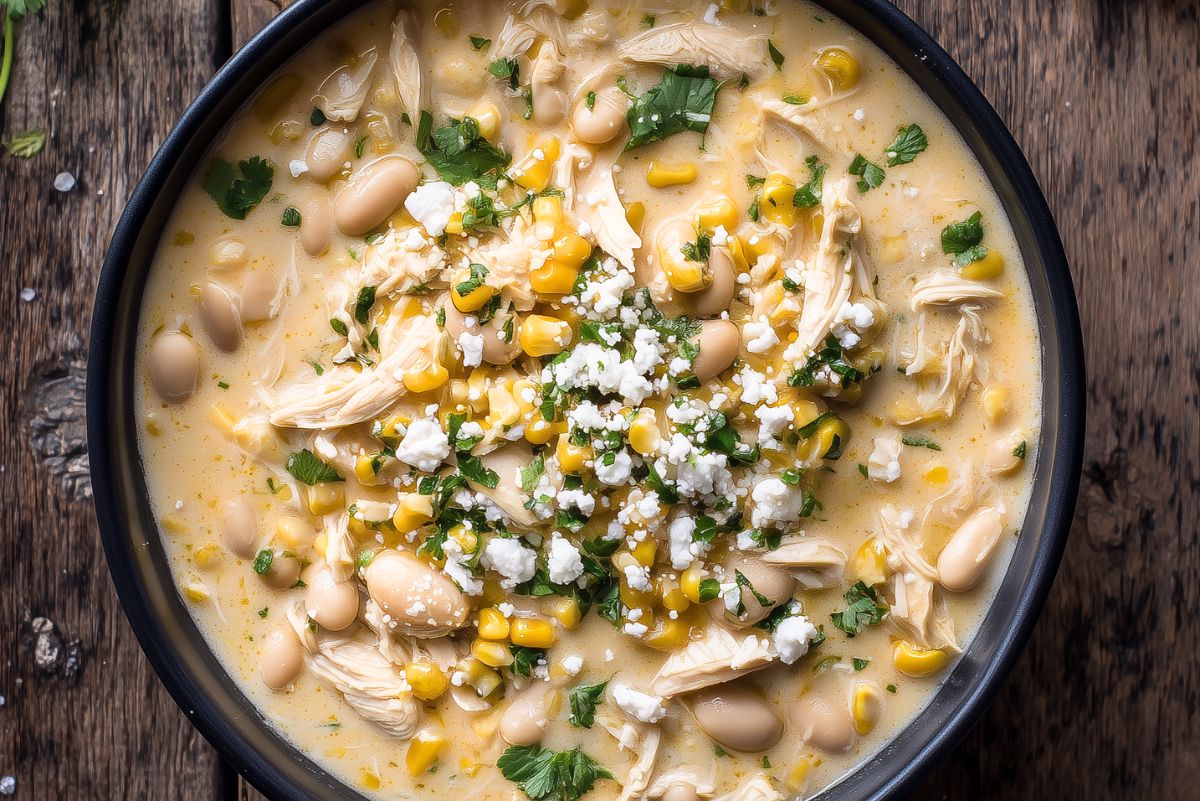 Overhead close-up of creamy Mexican street corn chicken soup with shredded chicken, corn, white beans, crumbled cheese, and chopped cilantro in a black bowl.