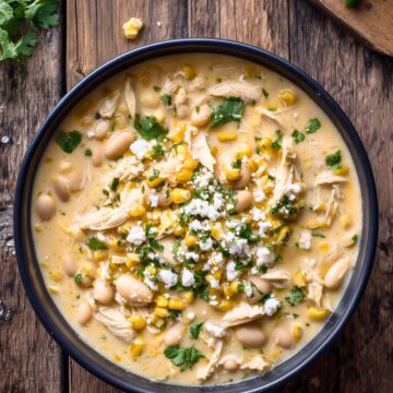 Creamy Mexican street corn chicken soup with shredded chicken, corn, white beans, crumbled cheese, and cilantro in a black bowl on a rustic wood table.