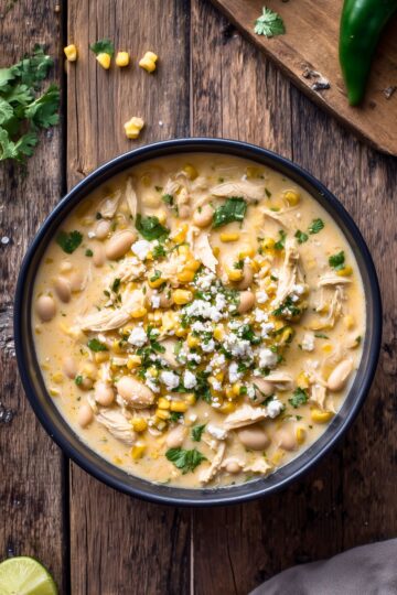 Creamy Mexican street corn chicken soup with shredded chicken, corn, white beans, crumbled cheese, and cilantro in a black bowl on a rustic wood table.