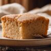 Old fashioned applesauce coffee cake with a moist, golden-brown crumb and lightly sugared top, served as a thick square slice on a rustic ceramic plate with a fork.