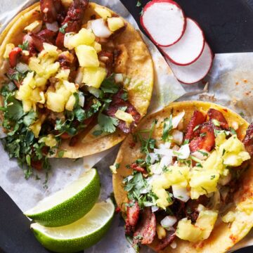 Pork tenderloin tacos in warm corn tortillas topped with fresh pineapple salsa, chopped cilantro, and white onion. Served on a black plate with lime wedges and sliced radishes.