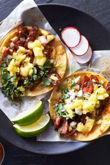 Pork tenderloin tacos in warm corn tortillas topped with fresh pineapple salsa, chopped cilantro, and white onion. Served on a black plate with lime wedges and sliced radishes.