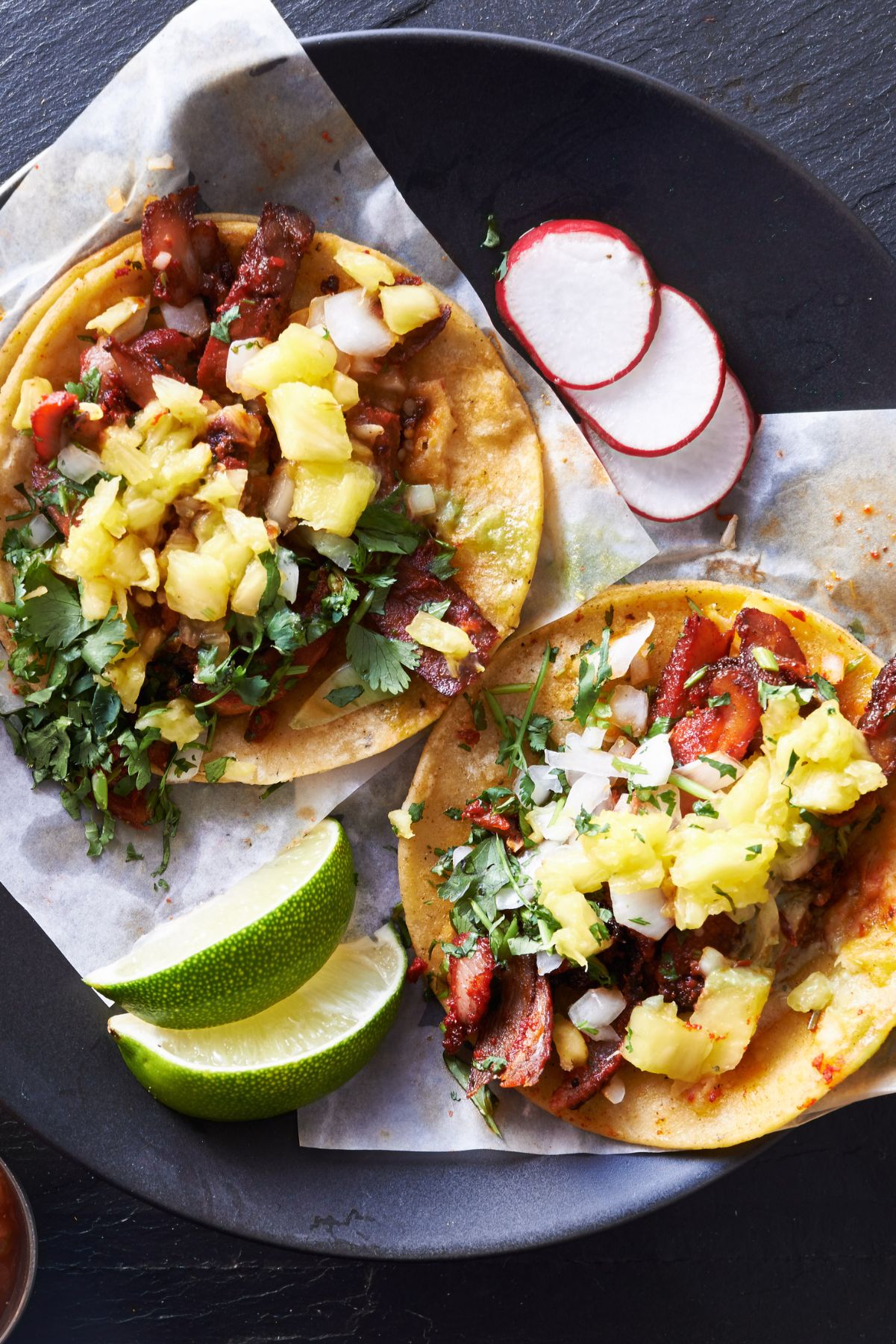 Pork tenderloin tacos in warm corn tortillas topped with fresh pineapple salsa, chopped cilantro, and white onion. Served on a black plate with lime wedges and sliced radishes.