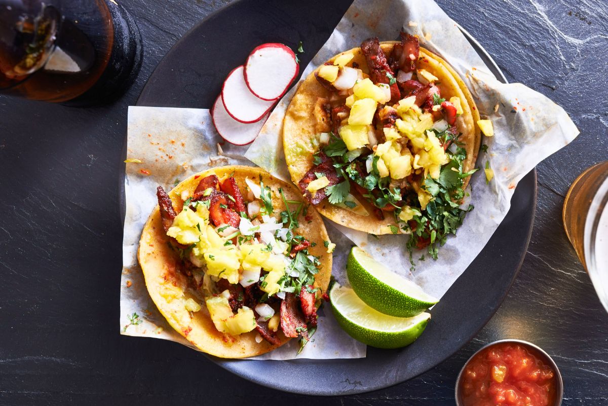 Pork tenderloin tacos topped with pineapple salsa, chopped cilantro, and white onion on corn tortillas. Served on a black plate with lime wedges, sliced radishes, and red salsa on the side.