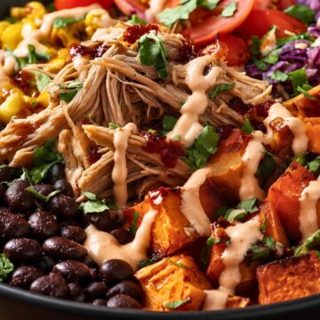 Pulled pork sweet potato bowl with roasted sweet potatoes, black beans, corn, tomatoes, red cabbage, cilantro, and creamy BBQ yogurt sauce in a black bowl.
