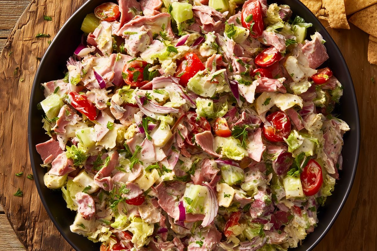 Overhead close-up of roast beef grinder chopped salad with lettuce, tomatoes, cucumber, red onion, cheese, herbs, and creamy dressing in a black bowl.
