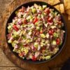 Overhead view of roast beef grinder chopped salad in a black bowl with lettuce, tomatoes, cucumber, red onion, cheese, herbs, and creamy dressing, served with pita chips on a rustic wooden table.