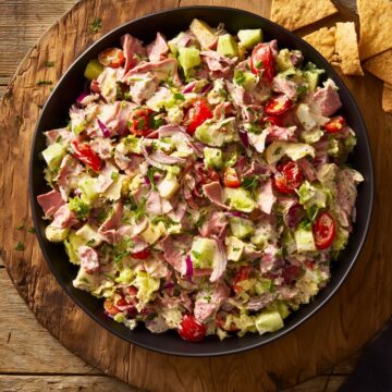 Overhead view of roast beef grinder chopped salad in a black bowl with lettuce, tomatoes, cucumber, red onion, cheese, herbs, and creamy dressing, served with pita chips on a rustic wooden table.