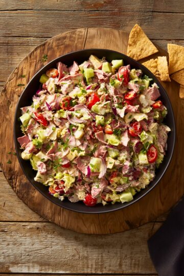 Overhead view of roast beef grinder chopped salad in a black bowl with lettuce, tomatoes, cucumber, red onion, cheese, herbs, and creamy dressing, served with pita chips on a rustic wooden table.