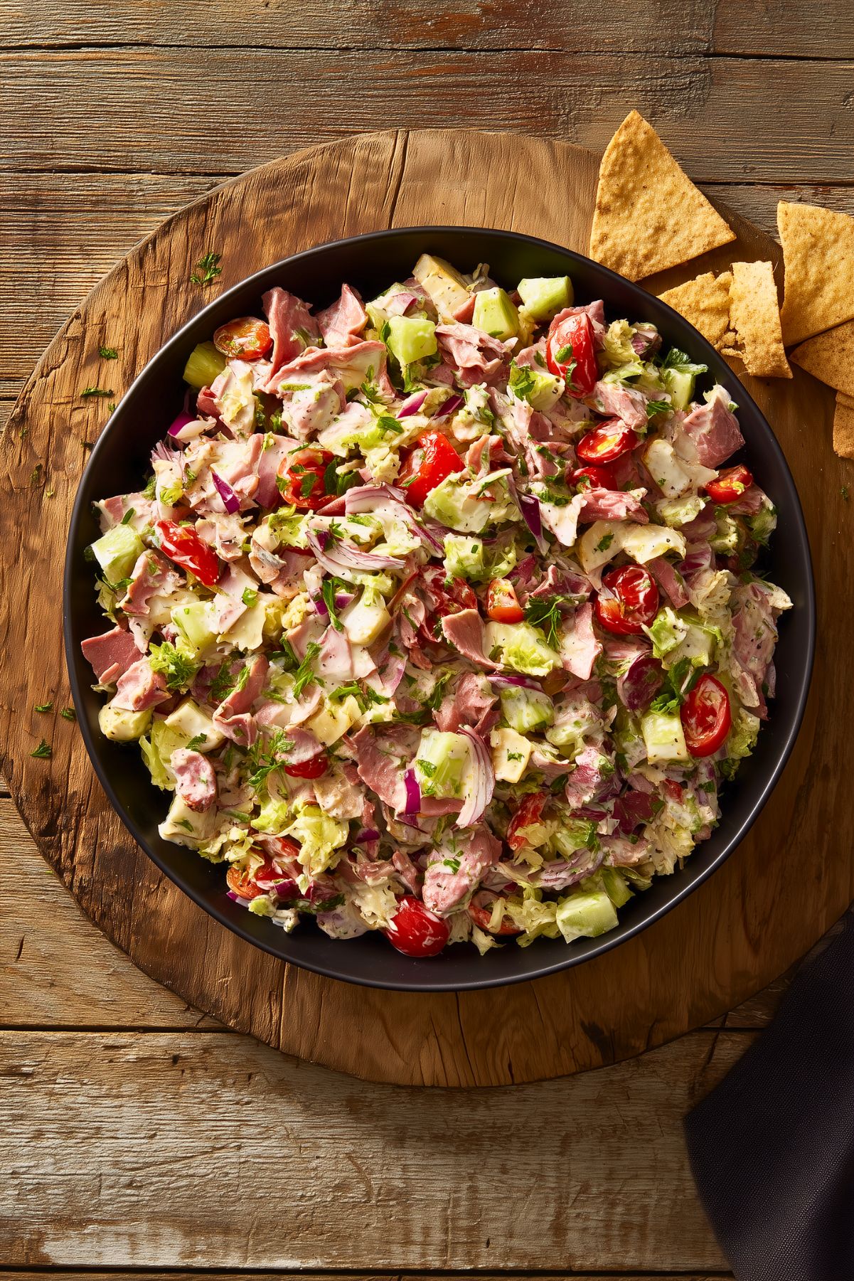 Overhead view of roast beef grinder chopped salad in a black bowl with lettuce, tomatoes, cucumber, red onion, cheese, herbs, and creamy dressing, served with pita chips on a rustic wooden table.