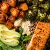 High protein salmon sweet potato bowl with seared salmon, roasted sweet potatoes and broccoli, quinoa, fresh spinach, avocado slices, and creamy dressing in a black bowl.