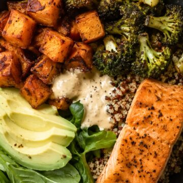 High protein salmon sweet potato bowl with seared salmon, roasted sweet potatoes and broccoli, quinoa, fresh spinach, avocado slices, and creamy dressing in a black bowl.