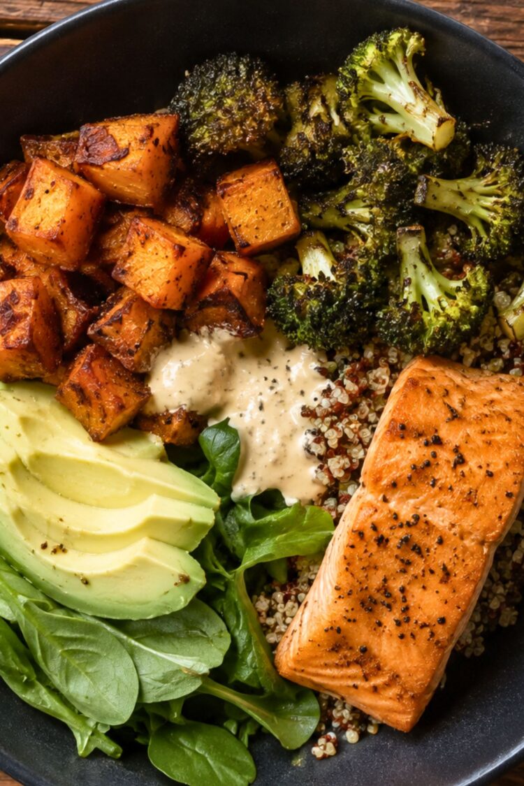 High protein salmon sweet potato bowl with seared salmon, roasted sweet potatoes and broccoli, quinoa, fresh spinach, avocado slices, and creamy dressing in a black bowl.