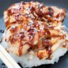 Skillet chicken teriyaki with tender sliced chicken glazed in glossy teriyaki sauce, served over steamed white rice and topped with sesame seeds on a dark plate with chopsticks.