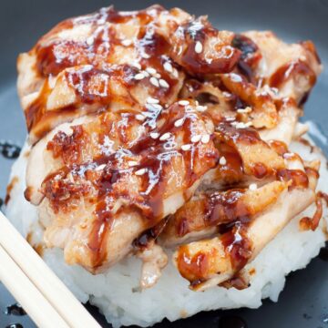 Skillet chicken teriyaki with tender sliced chicken glazed in glossy teriyaki sauce, served over steamed white rice and topped with sesame seeds on a dark plate with chopsticks.