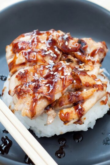 Skillet chicken teriyaki with tender sliced chicken glazed in glossy teriyaki sauce, served over steamed white rice and topped with sesame seeds on a dark plate with chopsticks.