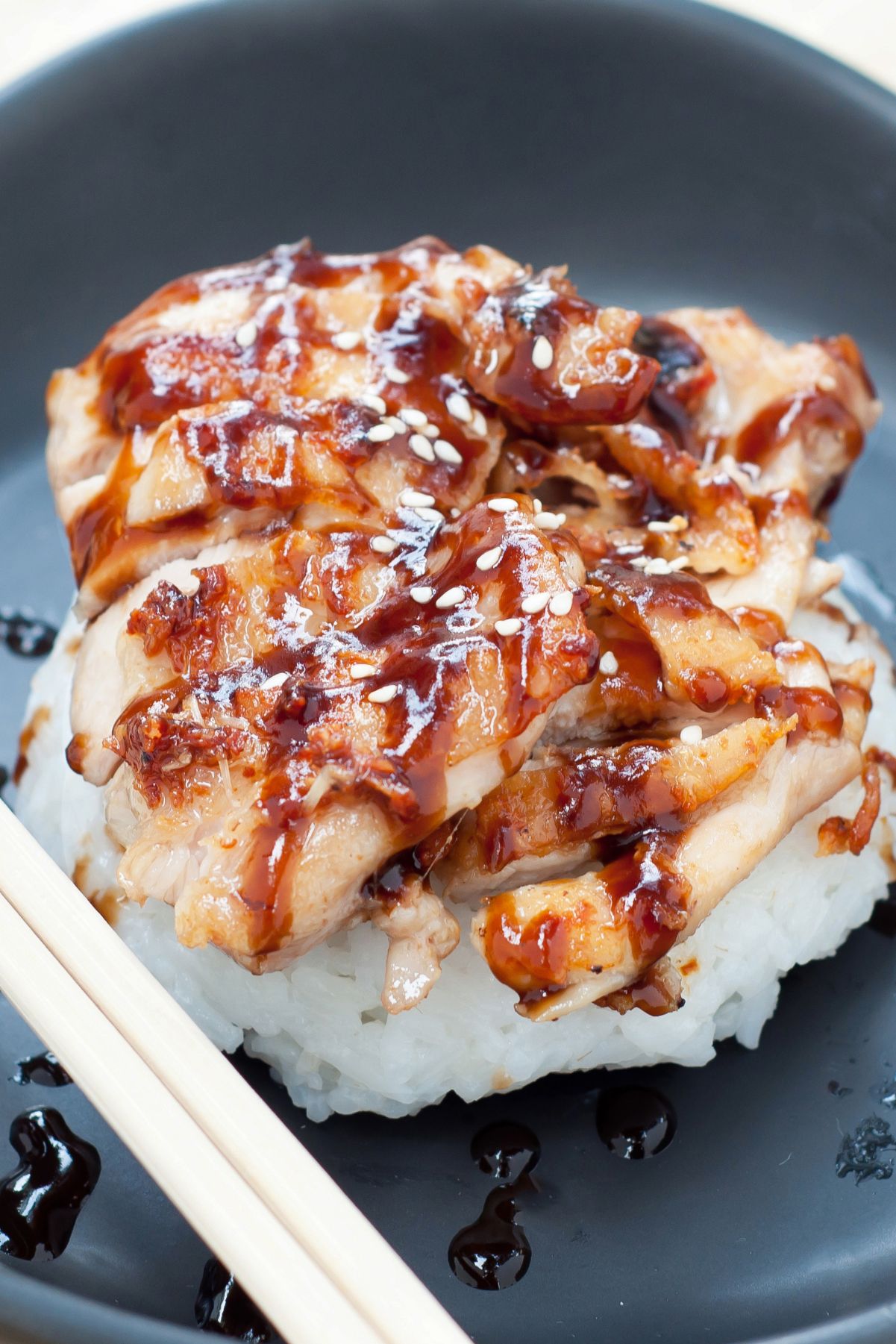 Skillet chicken teriyaki with tender sliced chicken glazed in glossy teriyaki sauce, served over steamed white rice and topped with sesame seeds on a dark plate with chopsticks.