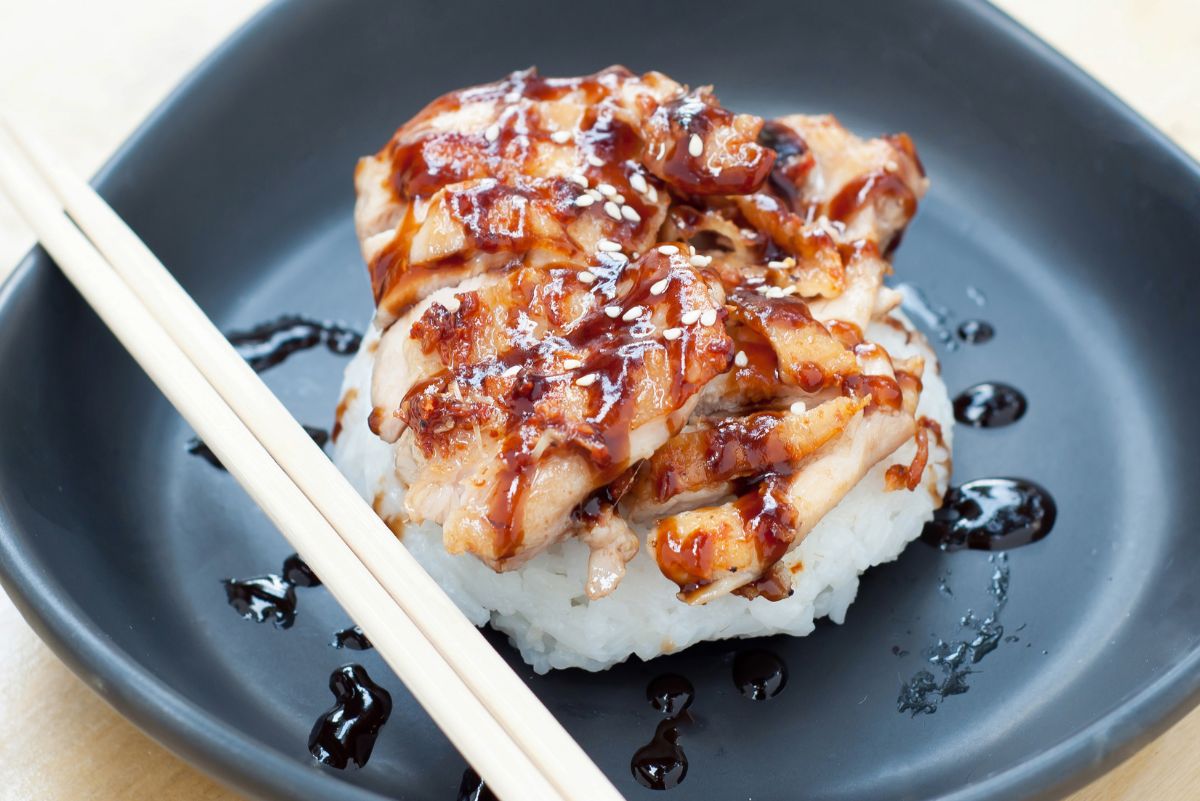 Skillet chicken teriyaki with sliced glazed chicken served over a mound of steamed white rice, topped with sesame seeds and extra teriyaki sauce on a dark plate with wooden chopsticks.