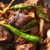South Indian lamb pepper fry with browned lamb pieces, green chilies, curry leaves, and sliced onions in a dark bowl.