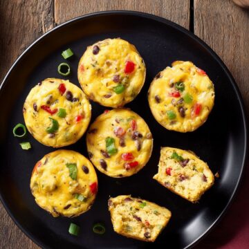 Overhead view of Southwest egg bites with black beans, red bell pepper, green onions, and cheese on a black plate, with two pieces cut open to show the interior.
