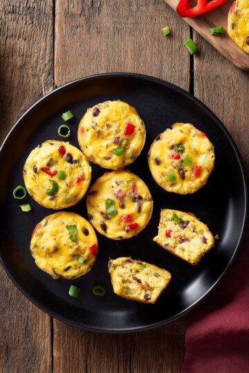 Overhead view of Southwest egg bites with black beans, red bell pepper, green onions, and cheese on a black plate, with two pieces cut open to show the interior.