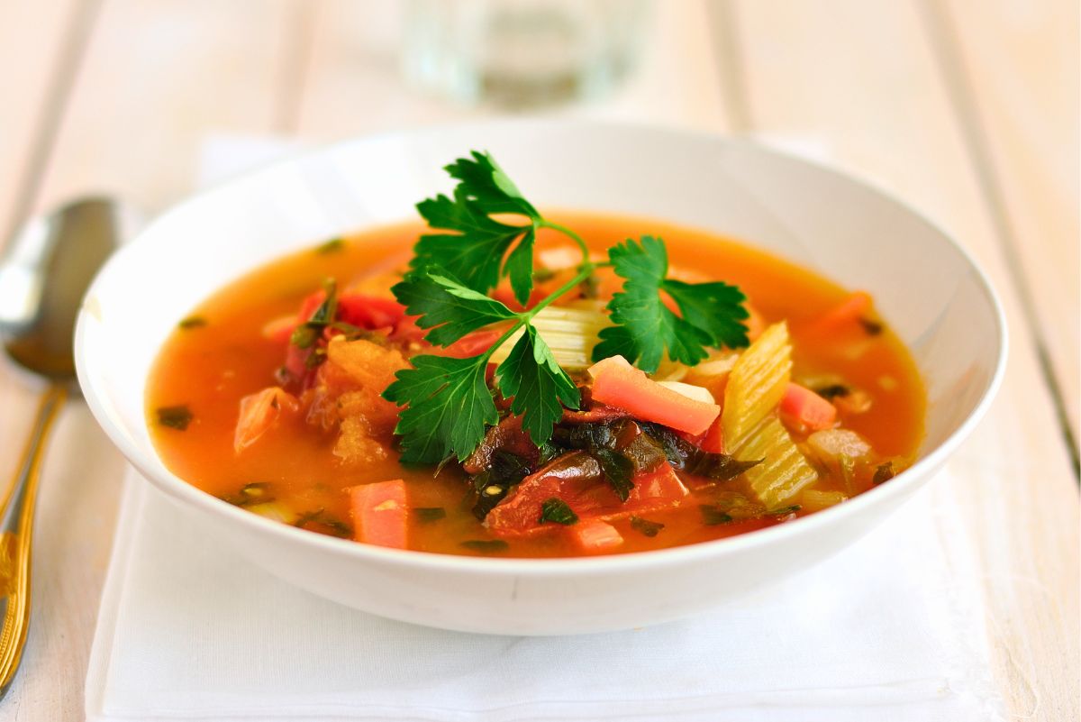 Spicy Tomato Trout and Vegetable Soup in a white bowl with trout, tomatoes, celery, red bell pepper, leafy greens, and fresh parsley in a tomato broth.