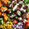 Steak sweet potato protein bowl with seared steak, roasted sweet potatoes, avocado, corn, cherry tomatoes, red onion, greens, and creamy dressing.