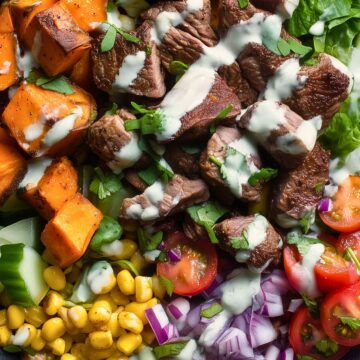Steak sweet potato protein bowl with seared steak, roasted sweet potatoes, avocado, corn, cherry tomatoes, red onion, greens, and creamy dressing.