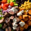 Overhead steak sweet potato protein bowl with sliced sirloin steak, roasted sweet potatoes, black beans, corn, tomatoes, green peppers, red onion, lettuce, cilantro, and creamy lime sauce in a black bowl on a rustic wood table.