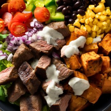 Overhead steak sweet potato protein bowl with sliced sirloin steak, roasted sweet potatoes, black beans, corn, tomatoes, green peppers, red onion, lettuce, cilantro, and creamy lime sauce in a black bowl on a rustic wood table.