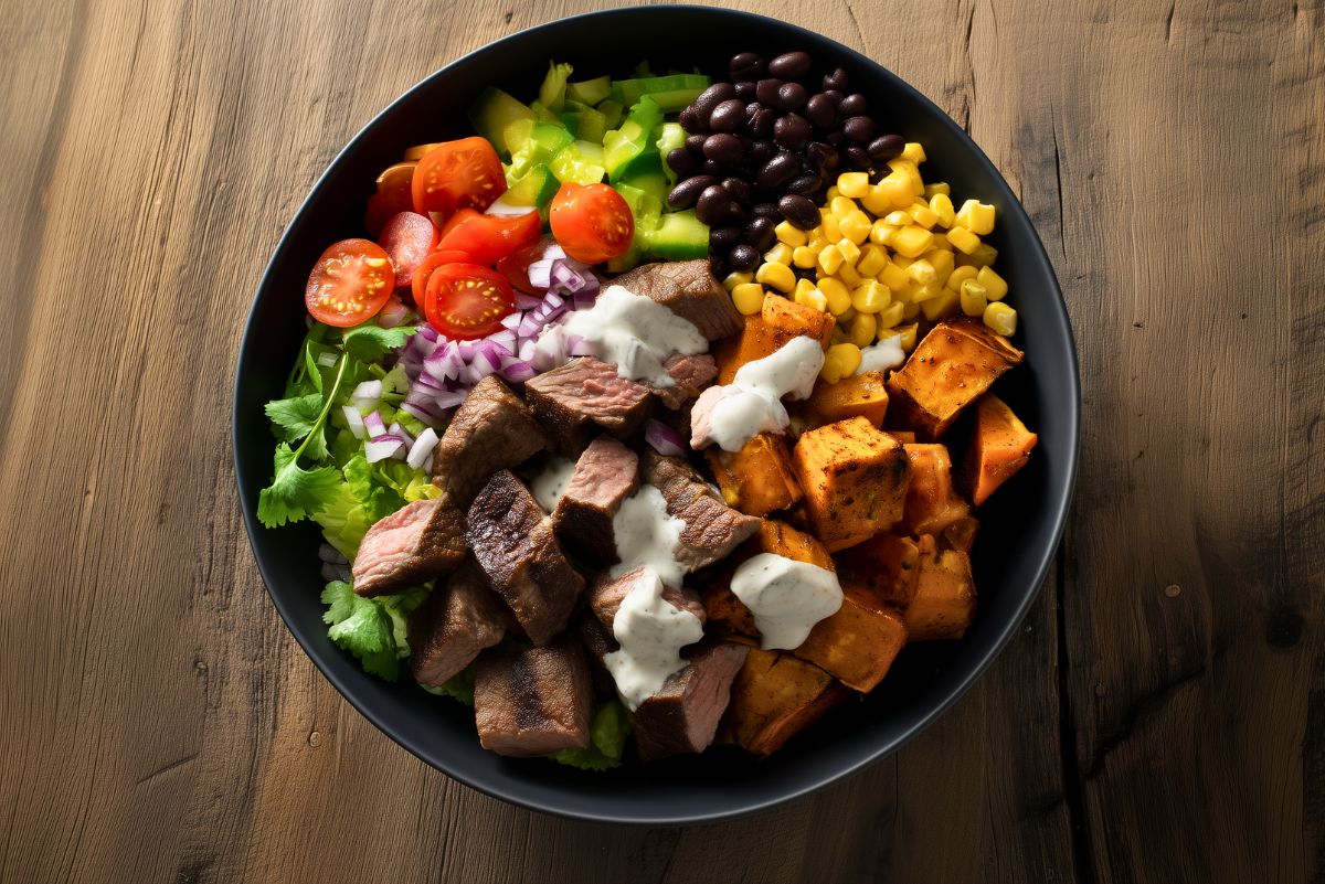 Overhead steak sweet potato protein bowl with sirloin steak, roasted sweet potatoes, black beans, corn, tomatoes, green peppers, red onion, lettuce, cilantro, and creamy lime sauce in a black bowl on a rustic wood table.