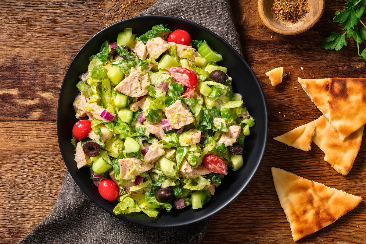 Turkey Grinder Chopped Salad with turkey, romaine lettuce, cucumber, cherry tomatoes, red onion, black olives, and creamy dressing in a black bowl, served with toasted pita chips on a rustic wooden table.
