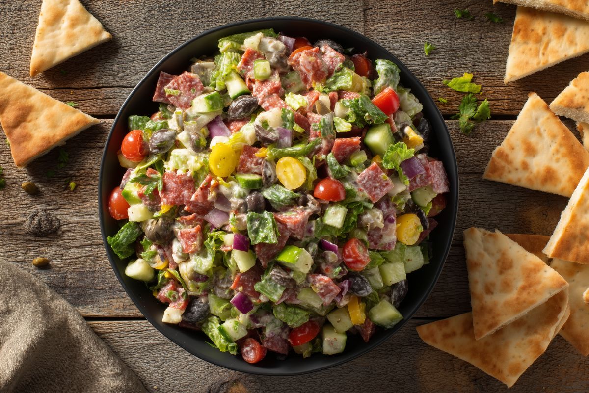 Turkey pepperoni grinder chopped salad with romaine, tomatoes, cucumber, red onion, banana peppers, black olives, provolone, and creamy Italian dressing in a black bowl surrounded by pita chips.