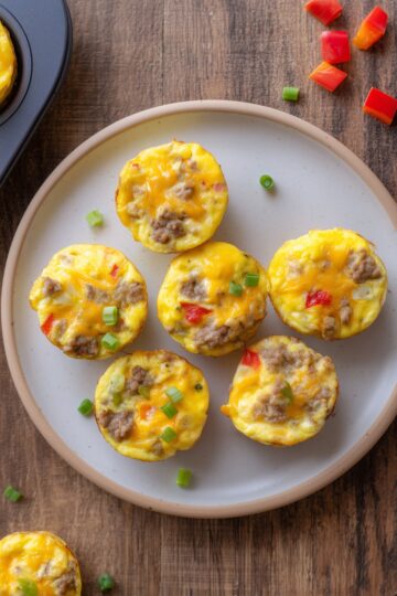 Six turkey sausage and Pepper Jack egg bites with melted cheese, red bell pepper, and green onions arranged on a plate over a rustic wood table.