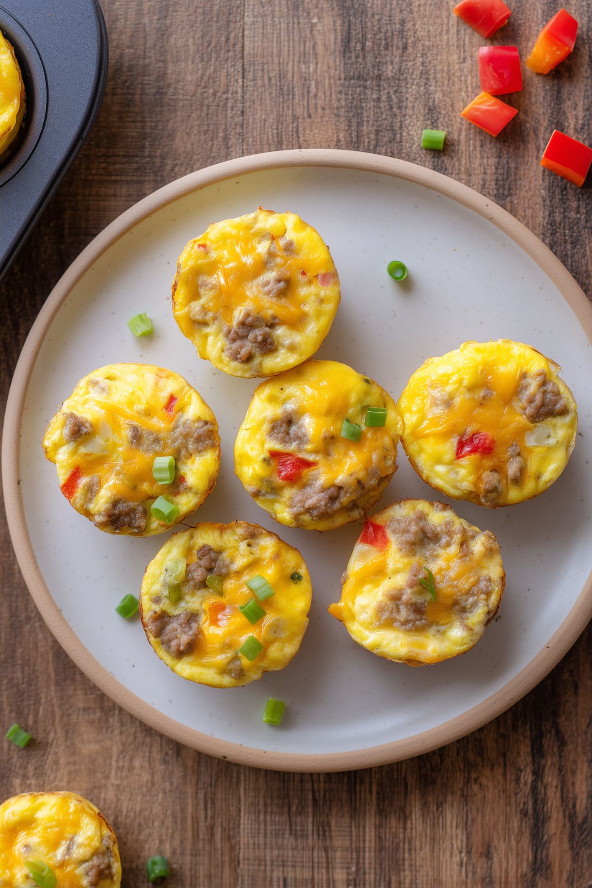 Six turkey sausage and Pepper Jack egg bites with melted cheese, red bell pepper, and green onions arranged on a plate over a rustic wood table.